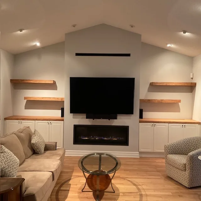 Vaulted-ceiling drywall fireplace with linear insert, floating wood shelves, and white cabinetry