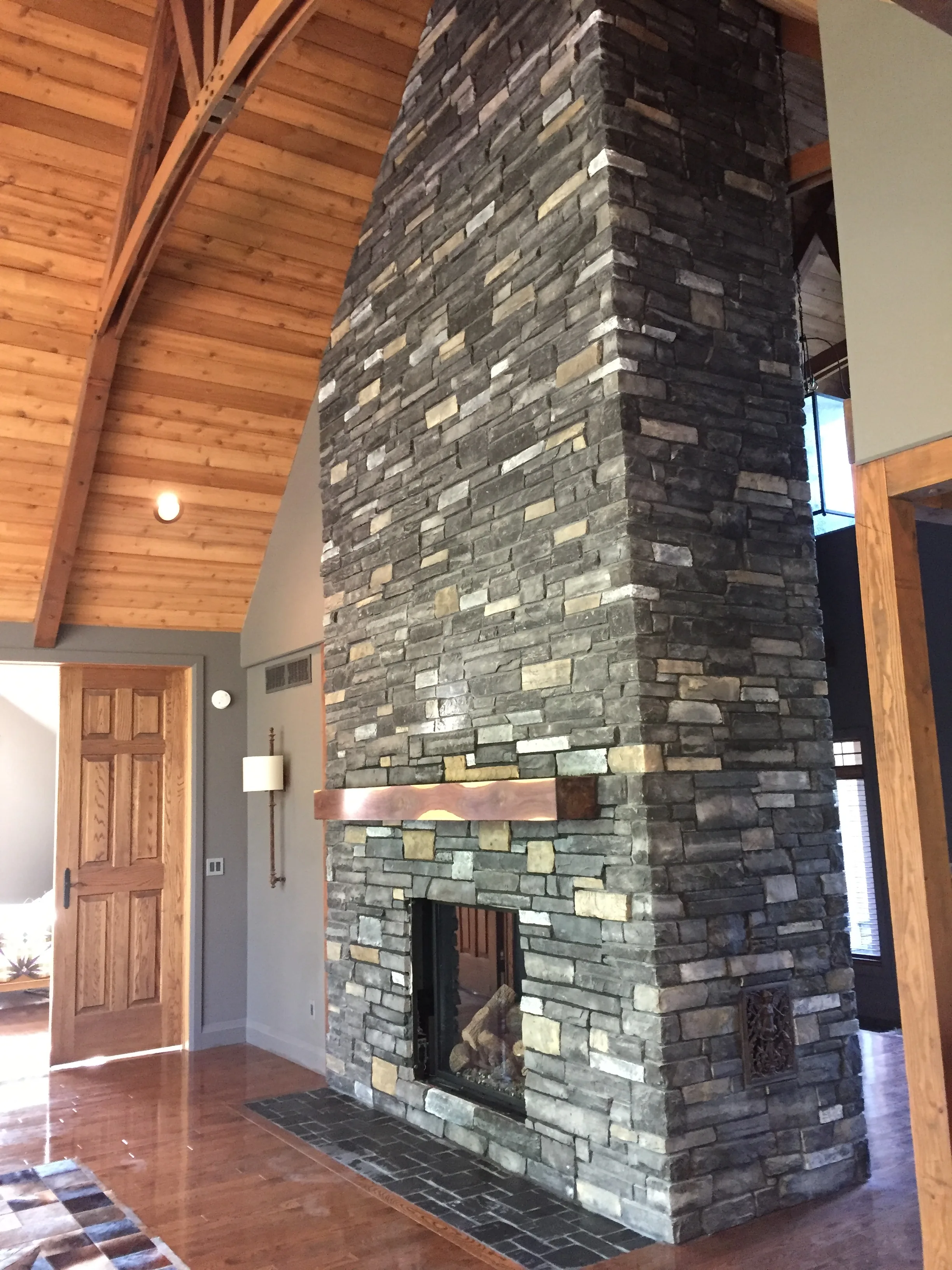 Two-story grey stacked-stone fireplace with copper accent bar in vaulted timber-ceiling great room