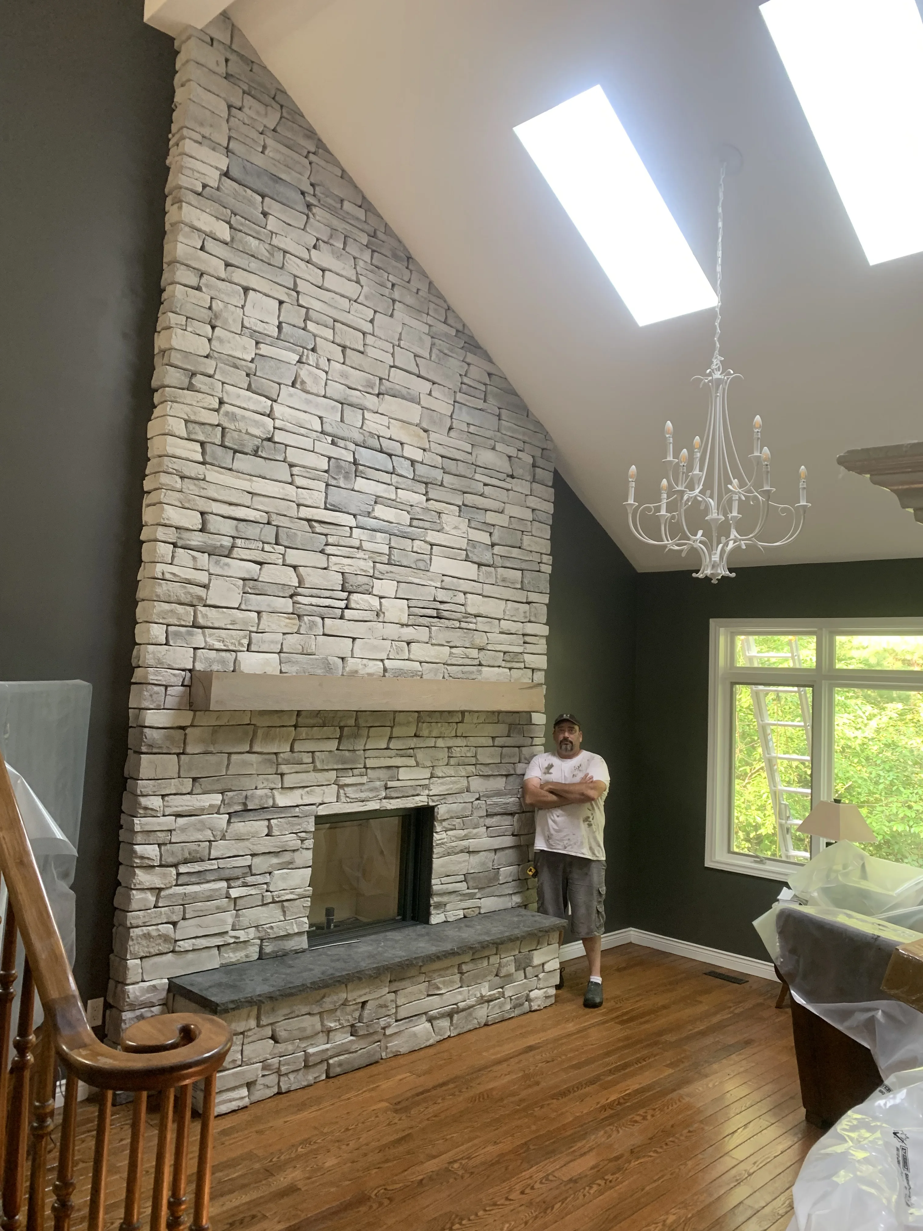 Jeff standing beside a two-story white stacked-stone fireplace he built, reaching up to a vaulted ceiling with skylights and a chandelier